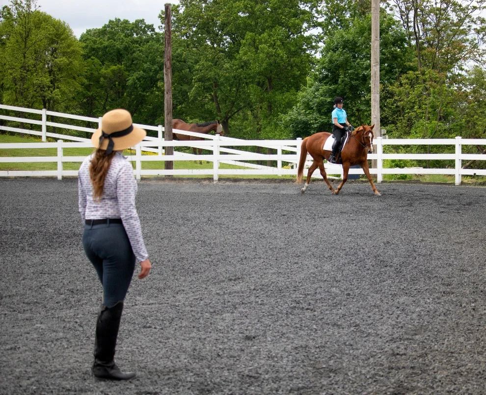 a horseback riding instructor trains a lesson student on the flat in a riding area. The student rides a chestnut horse